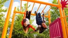 Two young people are hanging upside down from the yellow bars of a playground structure, embodying a carefree lifestyle. They are smiling and look playful. The background shows green trees and a blue sky with some clouds.