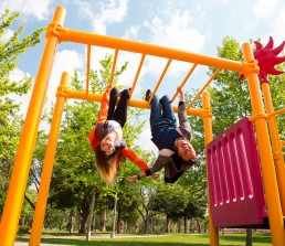 Two young people are hanging upside down from the yellow bars of a playground structure, embodying a carefree lifestyle. They are smiling and look playful. The background shows green trees and a blue sky with some clouds.