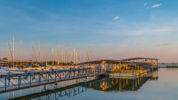 A peaceful marina with docked sailboats reflecting on calm water under a vibrant blue sky with light clouds during sunset. The docks stretch out from the shore, and the scene is bathed in warm, golden light, capturing a serene lifestyle by the water.