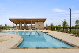 A sparkling outdoor swimming pool with lane markings is surrounded by a wide deck area and loungers. Alongside these amenities, behind the pool stands a wooden pavilion with stone supports, providing shaded seating. The area is fenced, and the sky is clear with a few scattered clouds.