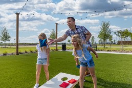 A man and two young girls are enjoying the amenities of a grassy field as they play a game of cornhole on a sunny day. The man tosses a blue bean bag while one girl prepares to throw, and the second girl stands ready to play. Trees surround them, with string lights adding charm above.